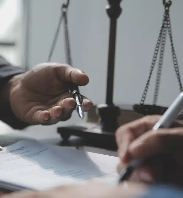 Close-up of two people’s hands, one passing a pen to the other, with a document being signed in the foreground and a blurred balance scale in the background, suggesting a legal or contract setting.