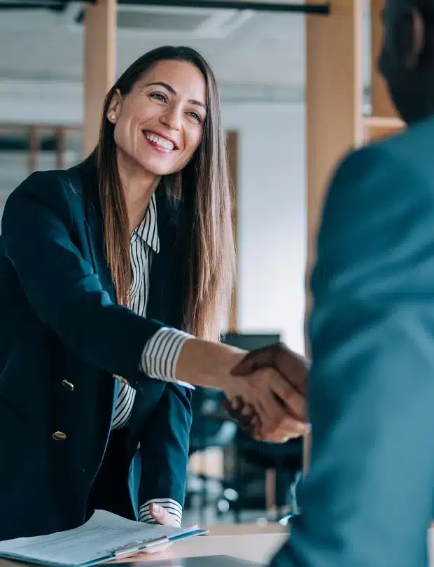 A smiling woman in business attire shakes hands with another person across a desk in an office, suggesting a successful meeting or interview.
