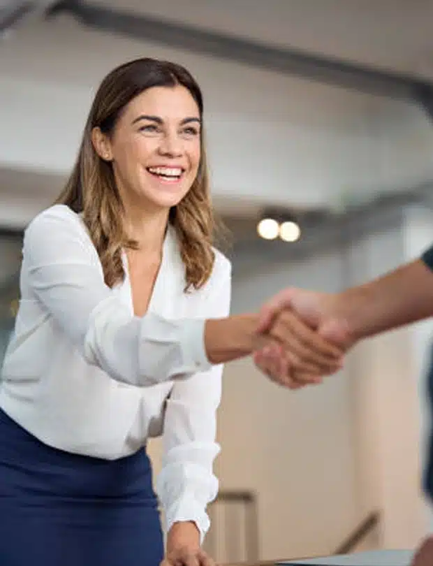 A smiling woman in a white blouse and blue skirt extends her hand to shake hands with another person, suggesting a friendly greeting or business agreement in a modern indoor setting.