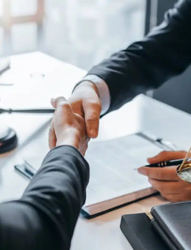Two people in business attire shake hands across a desk with an open notebook, pen, and legal documents, symbolizing agreement or partnership in a professional setting.
