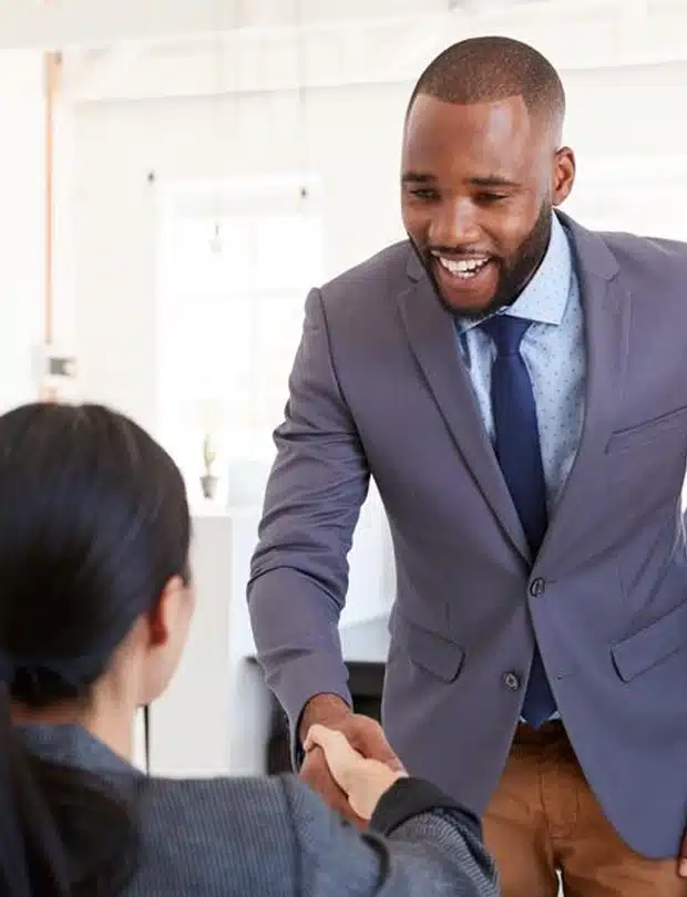 A smiling man in a suit leans forward to shake hands with a seated woman in an office setting.